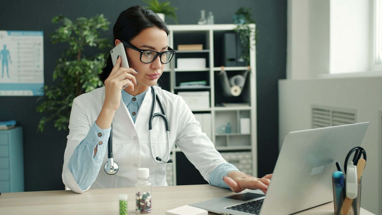 Médica ao telemóvel enquanto consulta a agenda no portátil do consultório